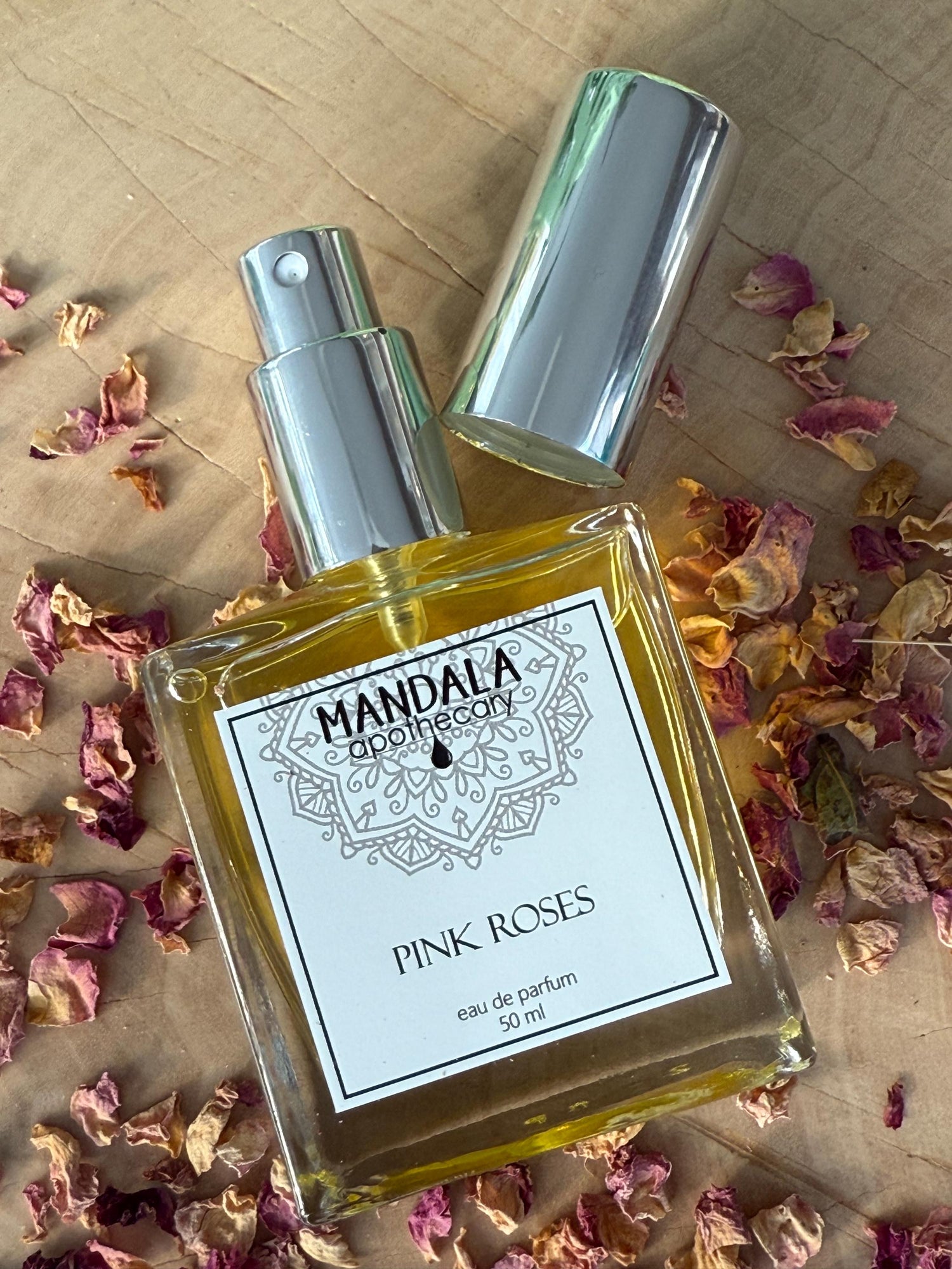 natural perfume spray made with essential oils of rose and citrus. Square spray bottle displayed on a block of wood surrounded by dried pink rose petals.